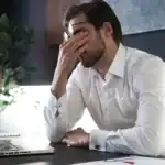 Frustrated man with hand on face, sitting at desk with laptop, representing workplace stress and mental health challenges; modern office environment for corporate wellbeing.