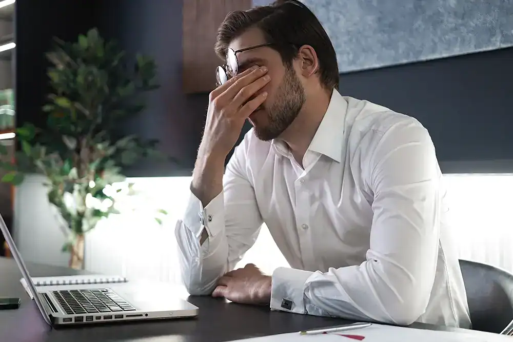 Frustrated man with hand on face, sitting at desk with laptop, representing workplace stress and mental health challenges; modern office environment for corporate wellbeing.